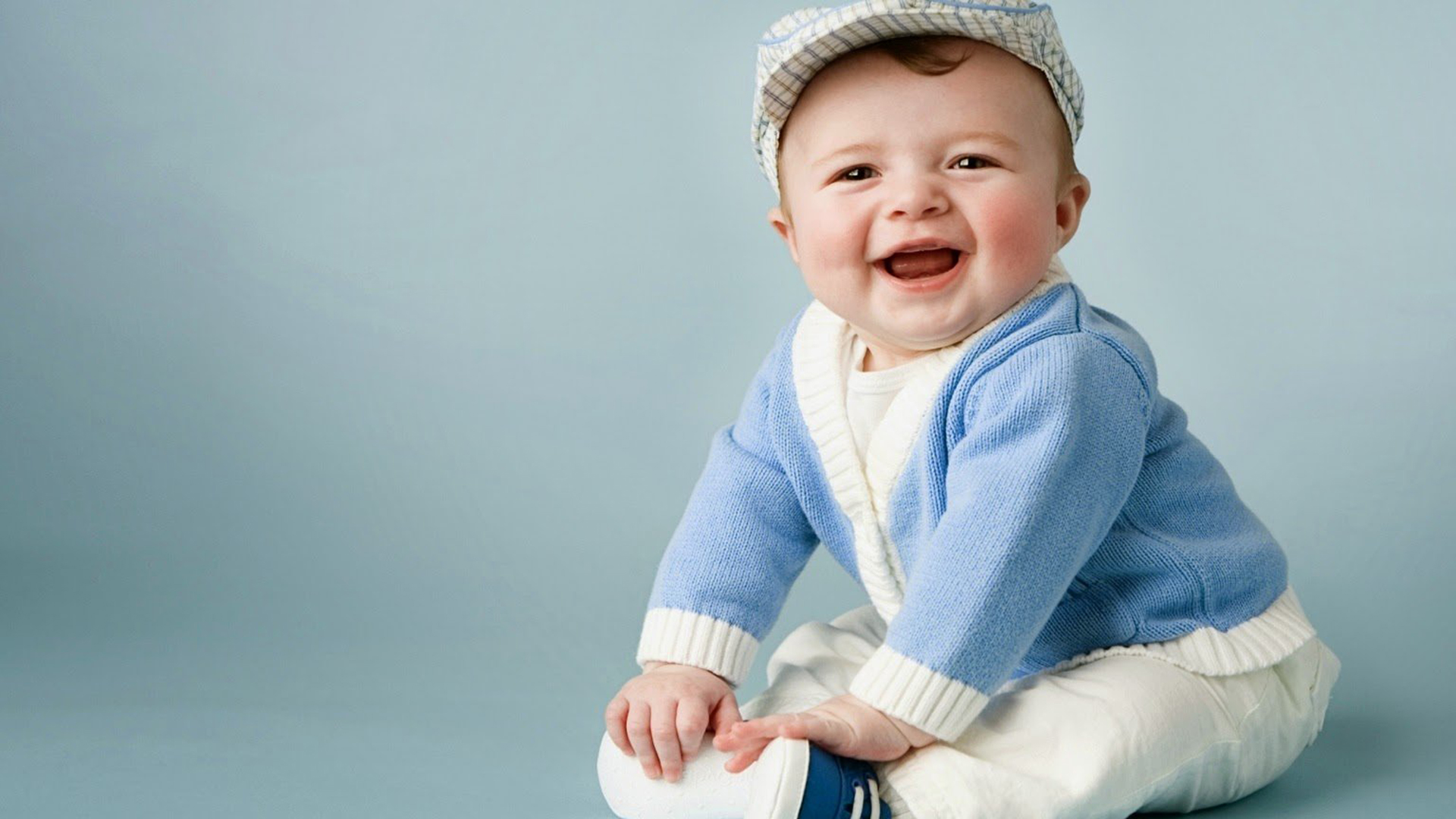 Smiley baby boy is sitting on floor wearing white dress and cap with blue woolen sweater in blue Wallpaper hd