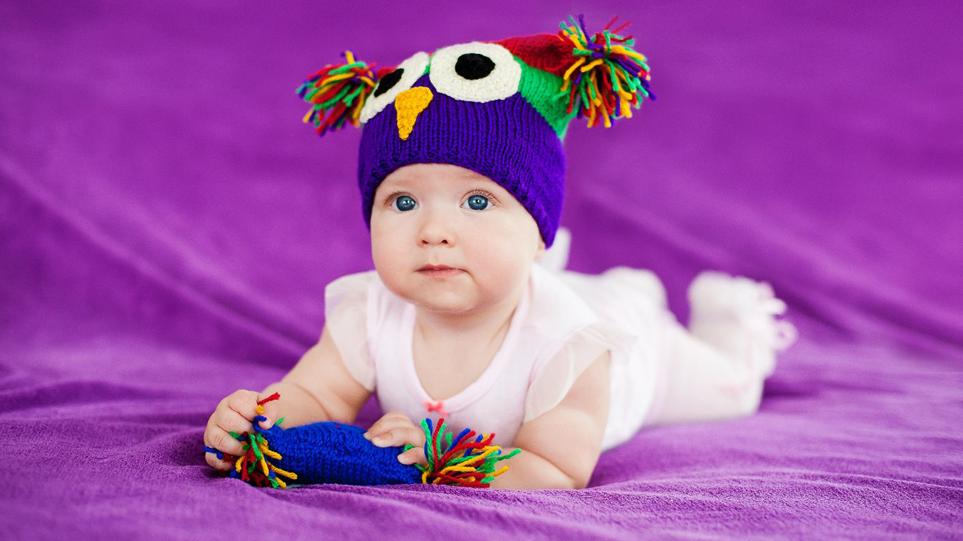 Girl Baby Is Lying Down On Soft Purple Cloth Wearing White Dress And Colorful Woolen Knitted Cap HD